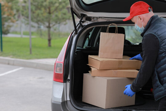 Young delivery man in protective mask, red cap and gloves near the car with boxes and packages, outdoors. Service coronavirus. Online shopping. mock up.