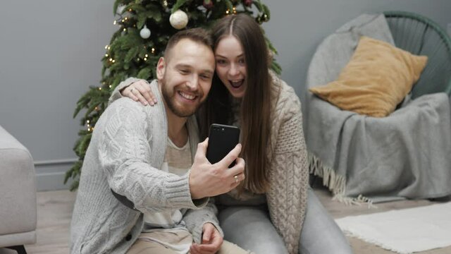 Happy cheerful man and woman in pullovers holding mobile phone and taking selfie or talking on video call with family on Christmas morning. Capture holiday memories on smartphone making selfie photo 