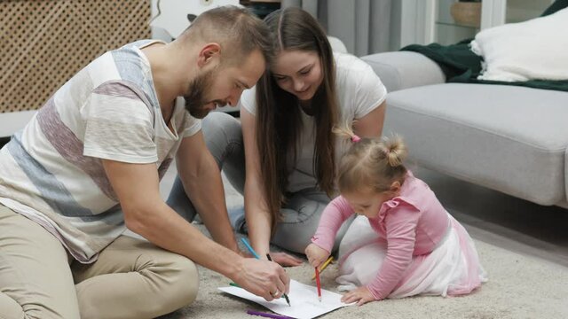 Happy family young parents drawing coloring picture with pencils helping cute child daughter enjoying talk play sitting on warm floor at home, mom dad and kid girl having fun in living room on leisure