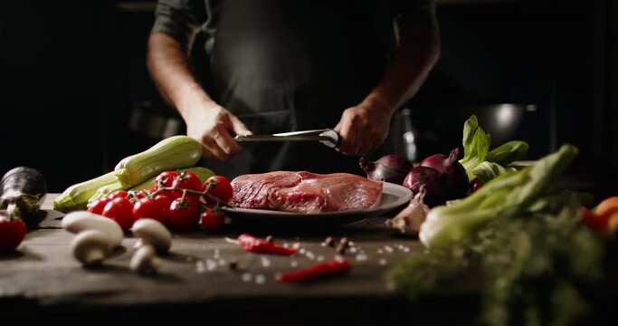 Chef sharpening his knife in front of kitchen table. Cooker preparing his tools before cutting raw piece of meat and various vegetables 4k footage