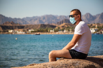 Fototapeta na wymiar tourist in a protective mask on vacation. man in a medical mask sits on the seashore