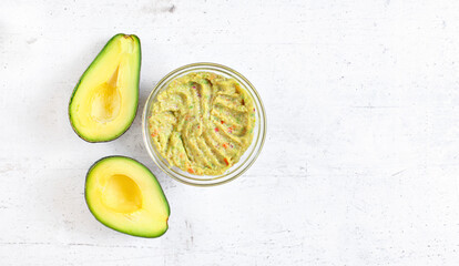 Two avocado halves, and glass bowl with prepared guacamole on white stone like desk, view from...