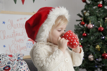 Adorable little girl in red Santa hat holding red mug at home in cozy holiday interior	
