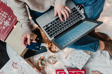 Christmas holiday online shopping. Gift boxes, lights. Festive season. Using laptop and credit card. Woman in cozy sweater and jeans uses laptop for shopping. Flat lay. Christmas boxes, ornaments.
