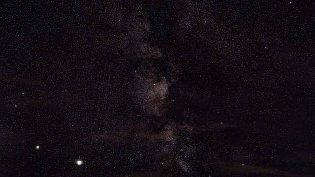 Night sky timelapse, many stars with satellites passing by, Milky way in region of Sagitta constellation visible, bright Saturn and Jupiter glowing lower part, clouds passing over
