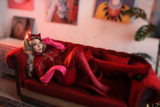 Sexy young woman in leather red dress with cat mask on the sofa