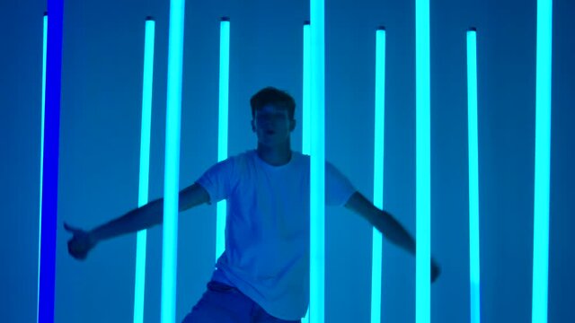 Young sporty man dancing hip hop against a background of bright blue neon lights. A silhouetted male dancer in casual youth clothing performs complex movements and jumps. Close up. Slow motion.