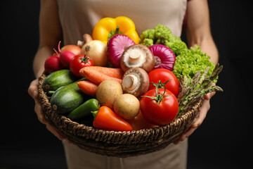 Woman holding wicker basket full of fresh vegetables on black background, closeup