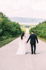 Newlyweds groom in a stylish black suit, the bride in a powdery dress, a photo session in nature, the road against the background of mountains and a reservoir in summer on a sunny day