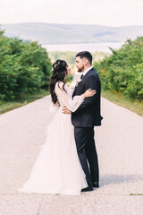 Newlyweds groom in a stylish black suit, the bride in a powdery dress, a photo session in nature, the road against the background of mountains and a reservoir in summer on a sunny day