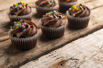 Chocolate cupcakes with sprinkling on rustic wooden background with yellow lights. Sweet dessert. Bakery concept. Elegant food. Sweets for coffee or tea.