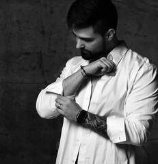Portrait of serious brutal handsome bearded man with tattoo on hand buttin up sleeve of shirt over...