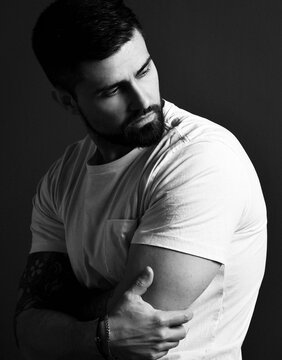 Portrait Of Serious Handsome Bearded Brutal Muscular Man With Tattoo On Hands In White T-shirt Embracing Hands And Looking Aside Over Dark Background. Hipster Stylish Look For Free Lifestyle Concept