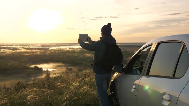 Driver blogger tourist shoots a video on modern gadget on vacation. Tourist with smartphone stopped at campsite by car, looking at sunset from mountain. Man traveling by car checks route in navigator