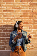 Vertical photo of young African American woman with headphones listening to music, singing and dancing fun on a brick wall.