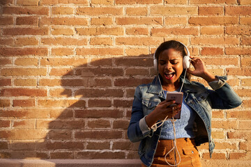 Portrait of young African American woman with headphones listening to music, singing loudly and excited on a brick wall.