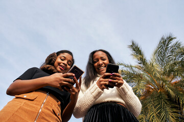 Low angle of two black girls using a mobile phone and laughing. One of them looks at the camera. People and technology concept with smartphone.