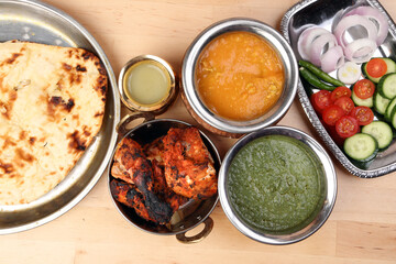 Spicy red Chicken grilled tikka tandoori nan bread green spinach curry yogurt sauce dal tomato cucumber onion salad set in metal stainless steel brass copper pot on light wood background