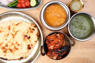 Spicy red Chicken grilled tikka tandoori nan bread green spinach curry yogurt sauce dal tomato cucumber onion salad set in metal stainless steel brass copper pot on light wood background