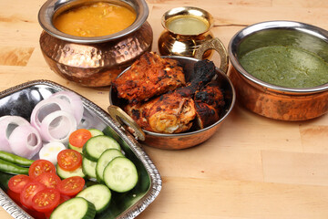 Spicy red Chicken grilled tikka tandoori nan bread green spinach curry yogurt sauce dal tomato cucumber onion salad set in metal stainless steel brass copper pot on light wood background