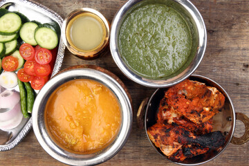 Spicy red Chicken grilled tikka tandoori nan bread green spinach curry yogurt sauce dal tomato cucumber onion salad set in metal stainless steel brass copper pot on rustic vintage wood background