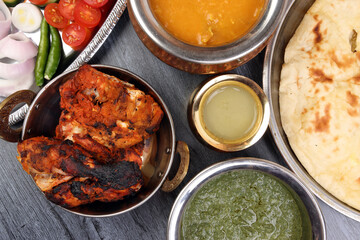 Spicy red Chicken grilled tikka tandoori nan bread green spinach curry yogurt sauce dal tomato cucumber onion salad set in metal stainless steel brass copper pot on black char wood background