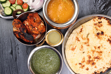 Spicy red Chicken grilled tikka tandoori nan bread green spinach curry yogurt sauce dal tomato cucumber onion salad set in metal stainless steel brass copper pot on black char wood background