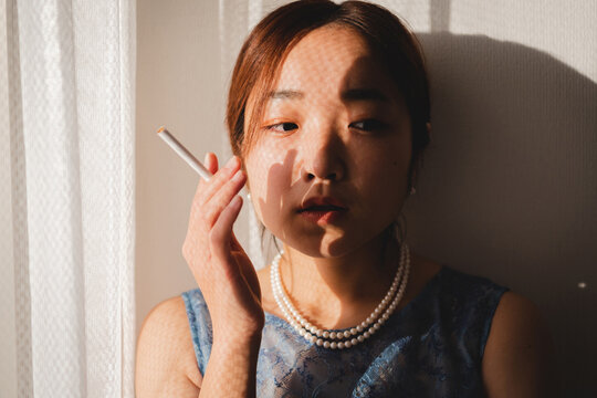 部屋の窓際でタバコを持つ青いドレスを着た若い女性