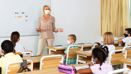 Young female teacher in protective face mask giving lesson to children in elementary school. New life reality during coronavirus pandemic