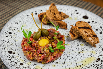Beef steak tartare with a quail egg yolk and capers on top, served with grilled croutons, on a white plate, close-up