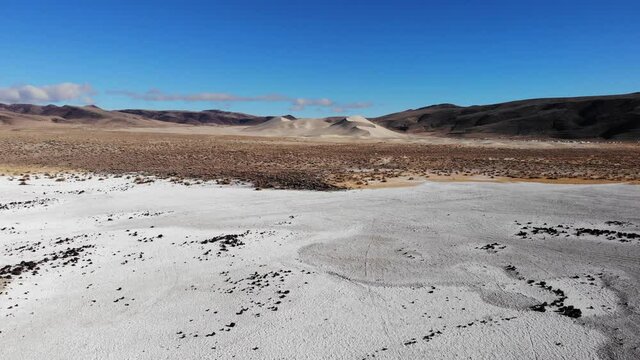 Flying Over White Salt Flats with Black Rocks and Sand Dune in Distance
