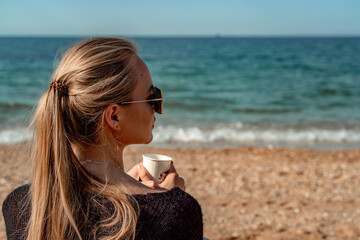 A beautiful blonde with long hair gathered in a ponytail sits on the beach drinking coffee and...
