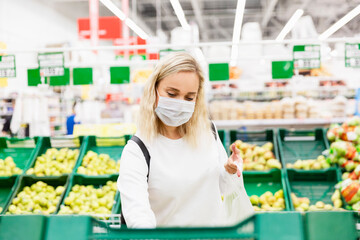 A young blonde woman in a medical mask buys fruits in a supermarket. Pandemic coronavirus.