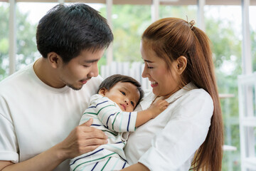 Obraz premium Asian father and mother are hugging 9 months baby son on sofa. They are smiling and warm touching to the baby with love in living room at home
