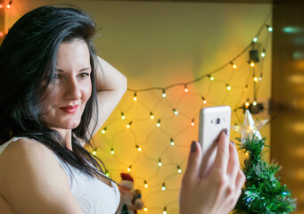 A beautiful brunette girl takes a selfie against the background of Christmas lights.