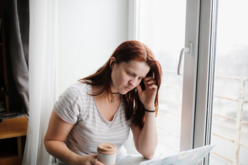 serious focused plus size caucasian woman by the window with phone and laptop, working remotely from home with poor connection. Concept of gadgets and communications during quarantine