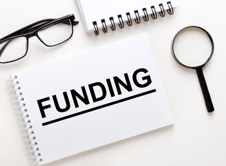 The word FUNDING is written in a white notebook on a light background near the notebook, black-framed glasses and a magnifying glass.
