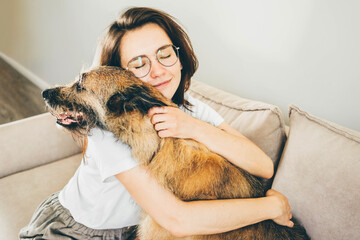 Positive brunette lady in white t-shirt with glasses hugs and strokes adorable old grey dog sitting...