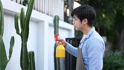 Side view of young Asian man using a water bottle to watering cactus at his garden.