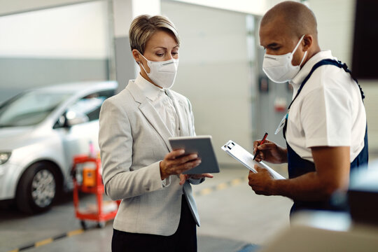 Businesswoman and African American mechanic wearing face masks and cooperating while using touchpad in a workshop.