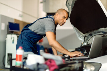 Black mechanic using laptop while running car diagnostic in a repair shop.