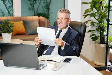 Positive senior businessman sitting at office desk and checking information in financial report