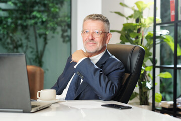 Portrait of positive aged entrepreneur in glasses sitting at office desk with cup of coffee and looking at camera