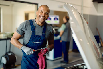 Happy black auto mechanic working at car workshop and looking at camera.