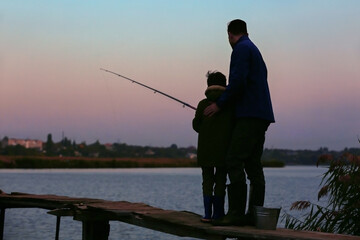 Silhouette of little boy and his father fishing on river in evening