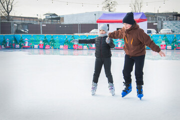 On a frosty winter day, brother and sister go ice skating on the ice rink. They are holding hands.