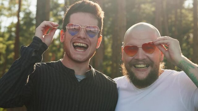 Homosexual couple smile nad laugh. Portrait of two mixed race gay men in love wearing sunglasses of heart shape smiling to camera. LGBTQI, Pride Event, LGBT Pride Month, Gay Pride Symbol.