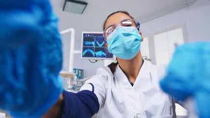 Patient point of view to dentist in protective mask holding tools examining person with toothache sitting on stomatological chair while nurse preparing tools for surgery.