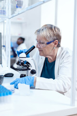 Senior scientist woman with protection glasses doing genetic investigation using microscope with african assistant in the background.. Elderly researcher carrying out scientific research in a sterile