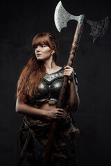 Obraz na płótnie Canvas Armed with huge axe and armoured woman viking with long brown hairs poses in dark background.